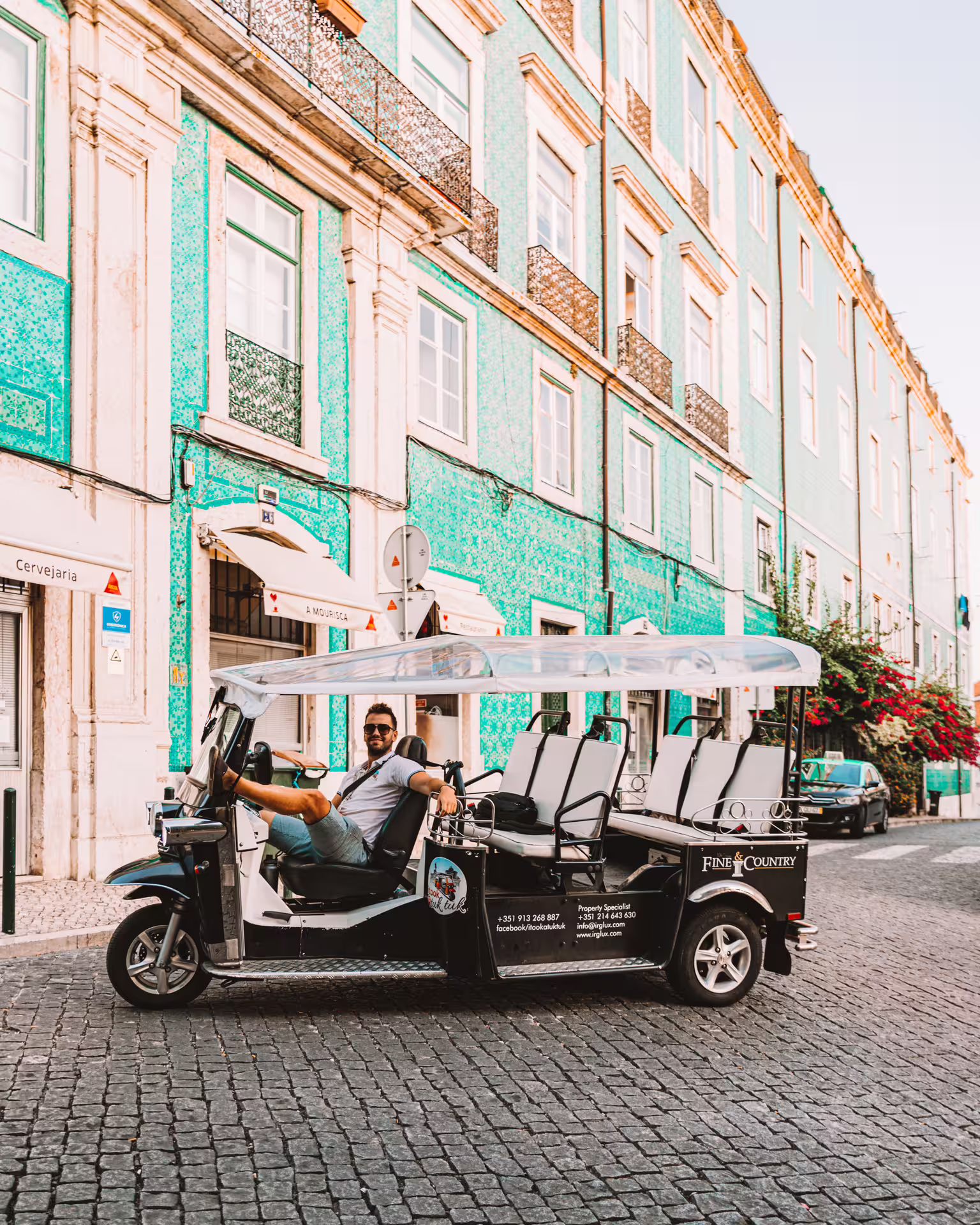 A friendly tuk-tuk driver smiles in front of Lisbon's colorful azulejo-tiled buildings, ready for a city tour adventure.