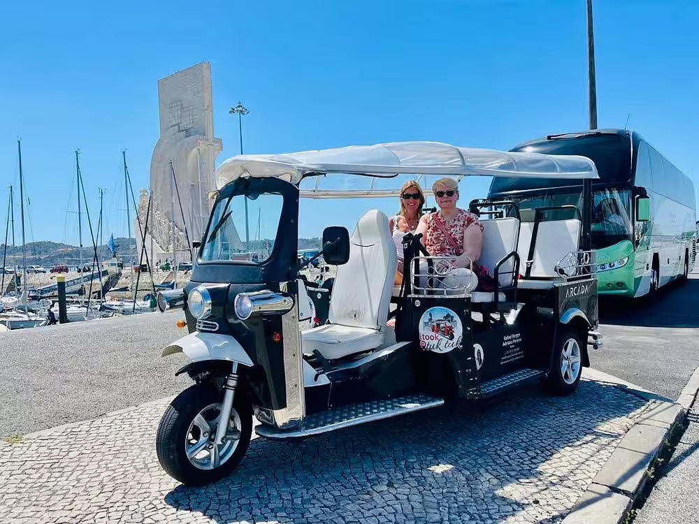 Tourists enjoying a sunny day on a tuk tuk ride along Lisbon's waterfront with iconic monument in the background.