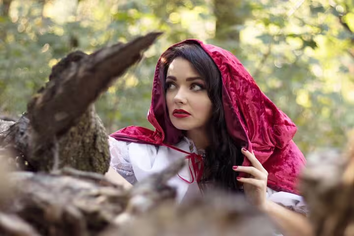 Woman dressed as Little Red Riding Hood in a forest, wearing a red velvet hood and white dress