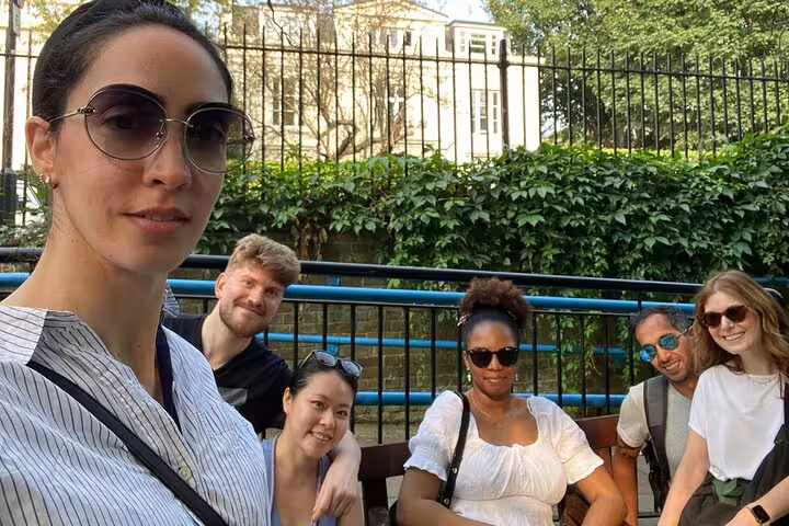 Group of six friends on a self-guided walking tour in London, sitting outdoors near iron fence and green hedge.