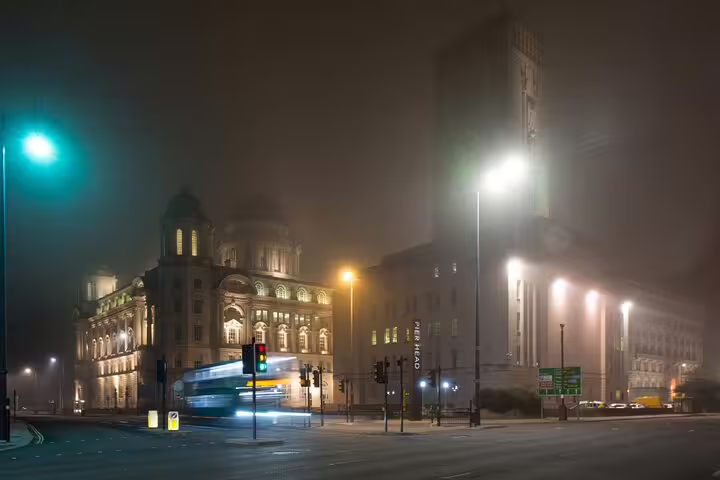 Foggy night at Liverpool Pier Head with a ghostly double-decker bus passing historic buildings