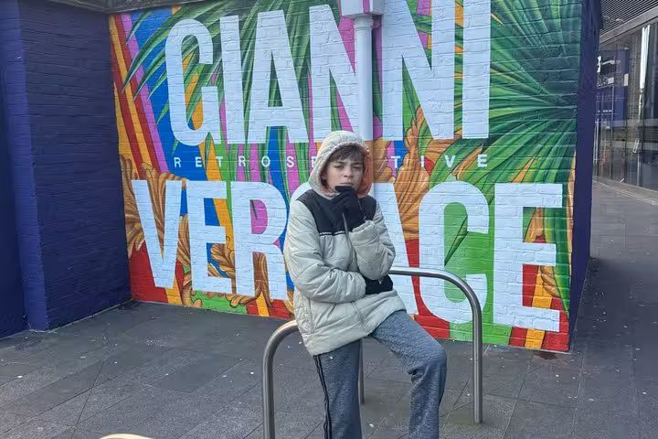 Person sitting on street railing in front of colorful Gianni Versace Retrospective mural in London