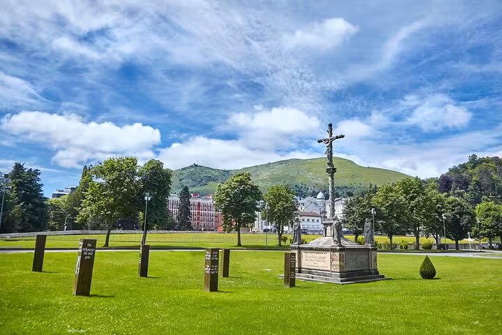 Scenic view of a cross monument in a lush green park in Lourdes, with hills and historic buildings in the background.