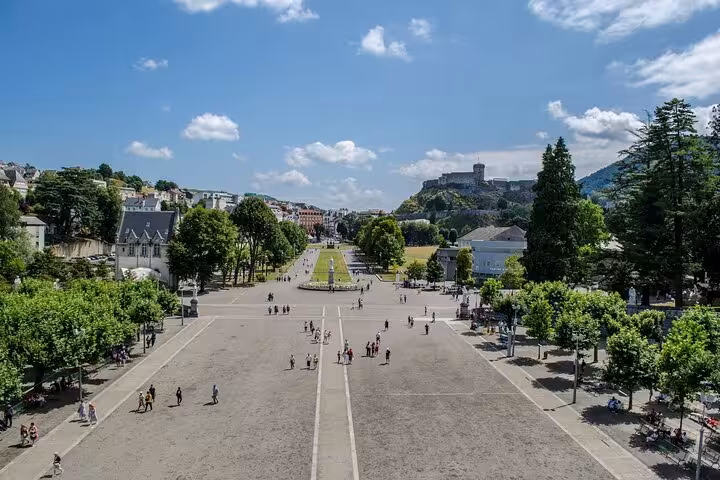 Panoramic view of a spacious plaza in Lourdes, lined with trees and leading towards the historic castle on a sunny day.