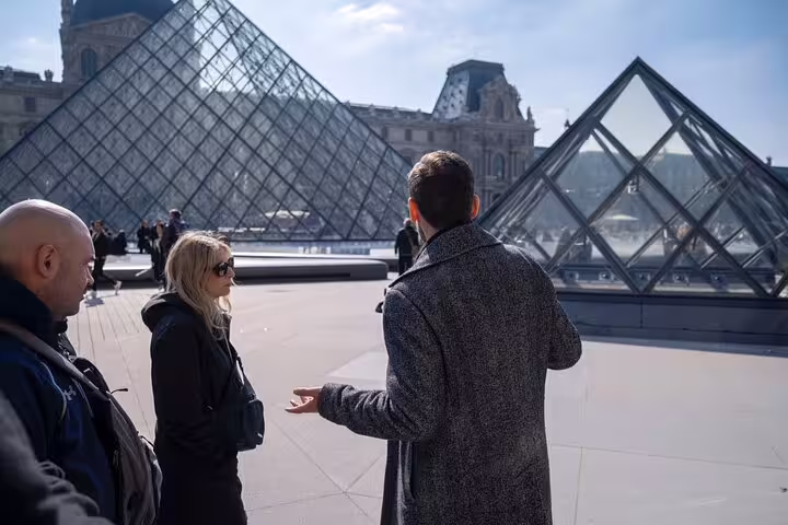 Tour guide explaining the history of the Louvre Museum to visitors near the iconic glass pyramid entrance.
