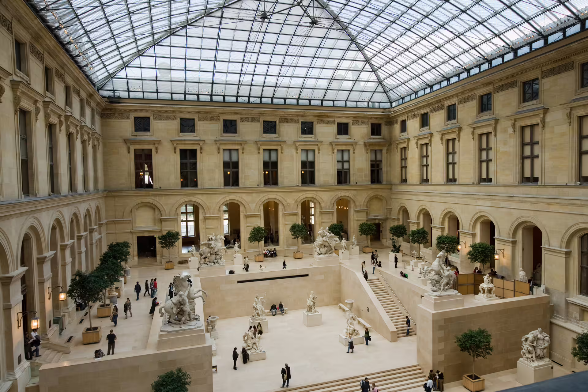 Grand glass-roofed sculpture hall at the Louvre Museum with marble statues and visitors
