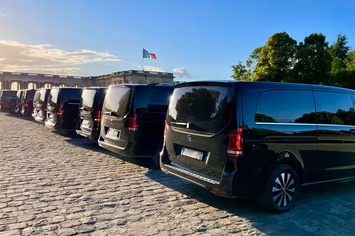 Luxury black vans lined up on cobblestone at Chantilly Chateau with French flag in background