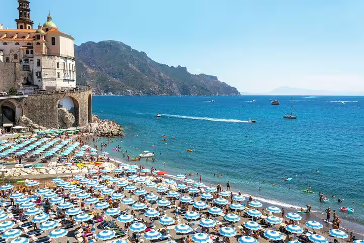 Crowded beach with blue parasols and historic seaside village, showcasing vibrant summer life on an Amalfi Coast catamaran cruise