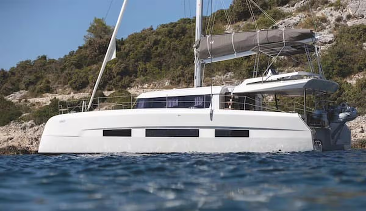 Modern luxury sailing catamaran anchored by a rocky green coastline on a weekly Mediterranean yacht charter cruise