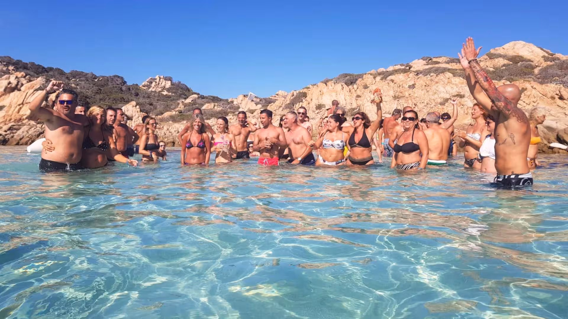 Excited group celebrating in the shallow waters of La Maddalena archipelago during a catamaran tour adventure.