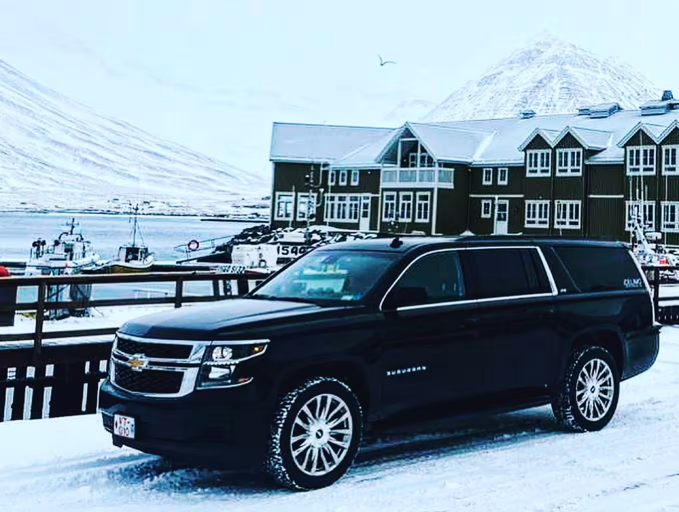 Black Chevrolet Suburban SUV parked in snowy Akureyri harbor with snow-capped mountains in background