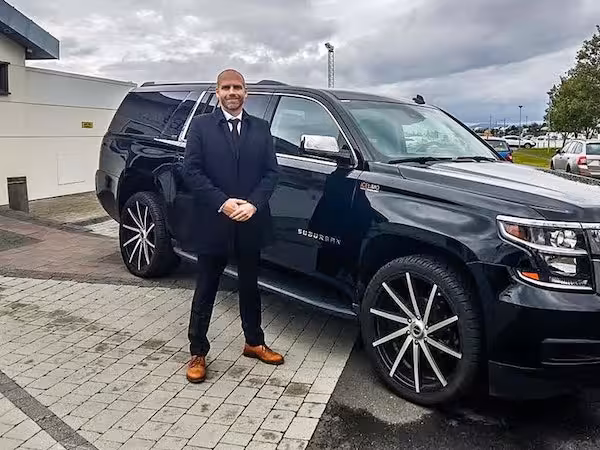 Professional driver in black suit standing beside a luxury black Chevrolet Suburban SUV on paved lot