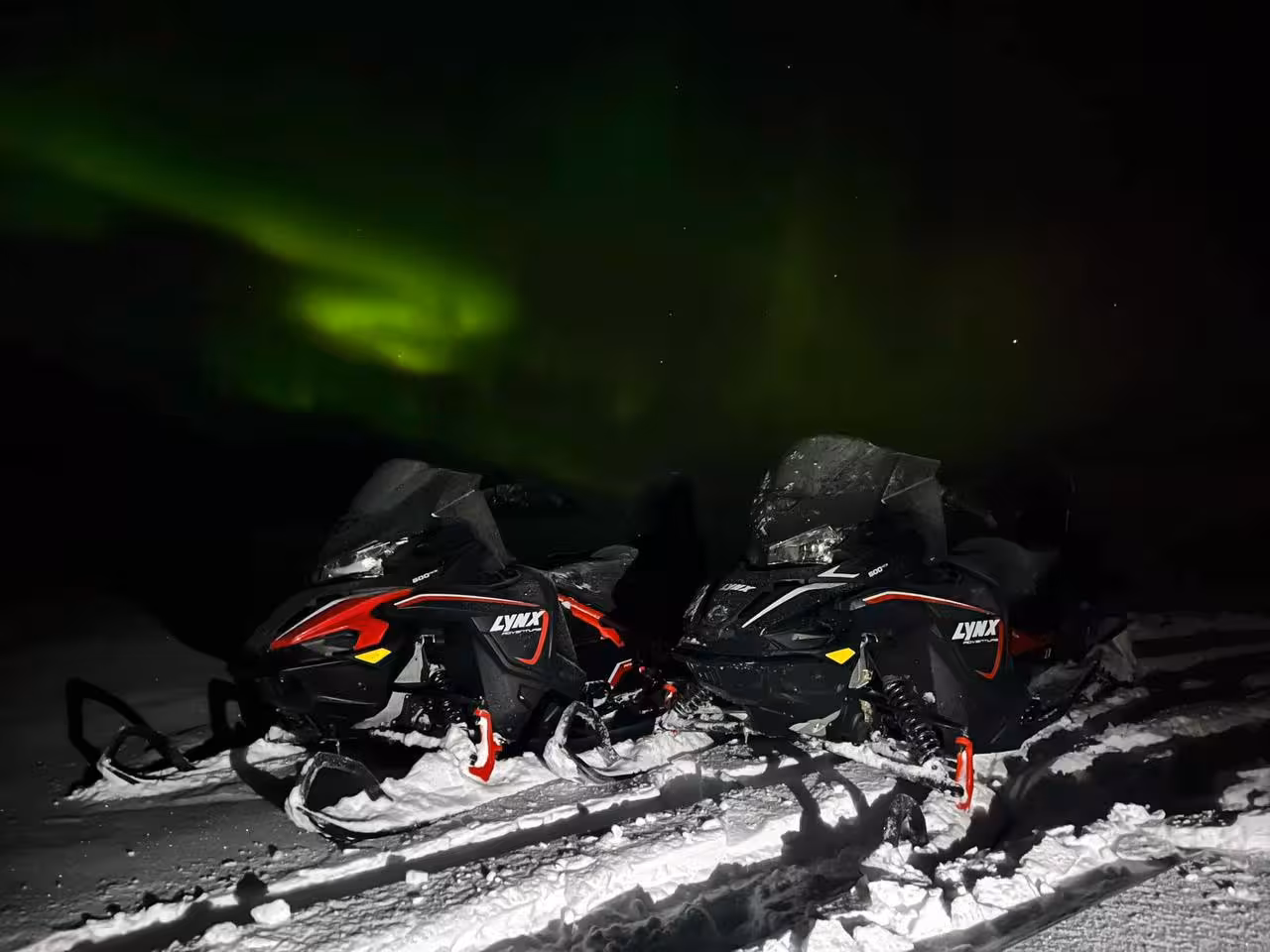 Two Lynx snowmobiles parked in snow under vivid green northern lights in Iceland highlands at night