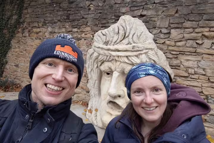 Two smiling tourists taking a selfie with a large stone face sculpture against a stone wall in Lyon