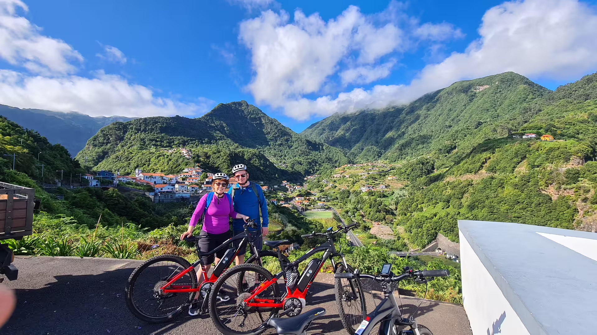 Couple enjoying a scenic e-bike tour on Madeira's lush north coast, with panoramic views of verdant mountains and villages.