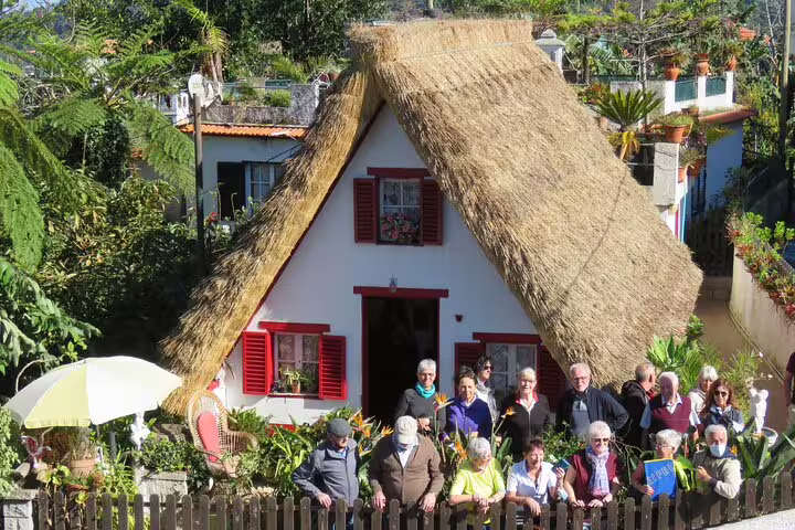 Tourists enjoy a full day guided tour in Madeira, visiting a traditional Santana house with a thatched roof and vibrant gardens.