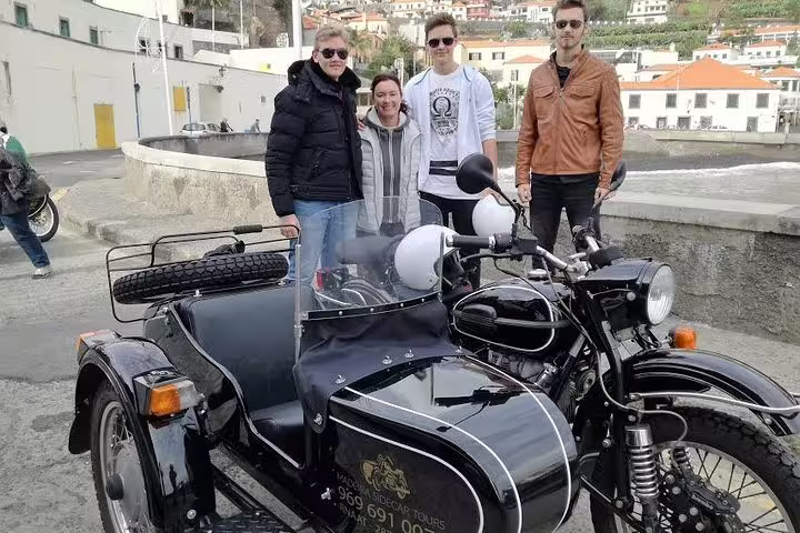 Group enjoying Madeira Old Road West sidecar tour, posing with vintage motorcycle, scenic backdrop of traditional architecture.
