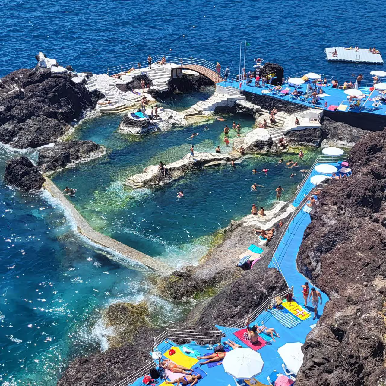 Aerial view of natural volcanic pools in Madeira with tourists enjoying the sun and crystal-clear Atlantic waters on a private tour.
