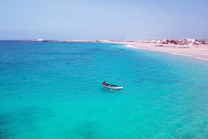 A small boat floats on the turquoise waters of Maio Island's coastline, with a view of the sandy beach and distant harbor.