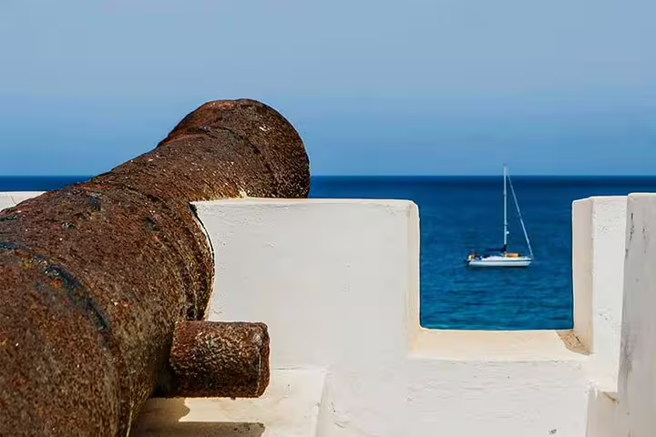 Rustic cannon overlooking the serene blue ocean with a sailboat in the distance on Maio Island, a historic tour highlight.