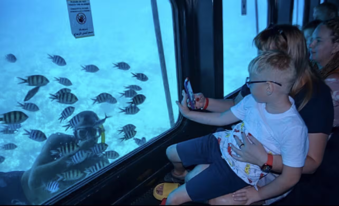 Family watching colorful fish through semi submarine window in Makadi Bay, child taking photo with smartphone