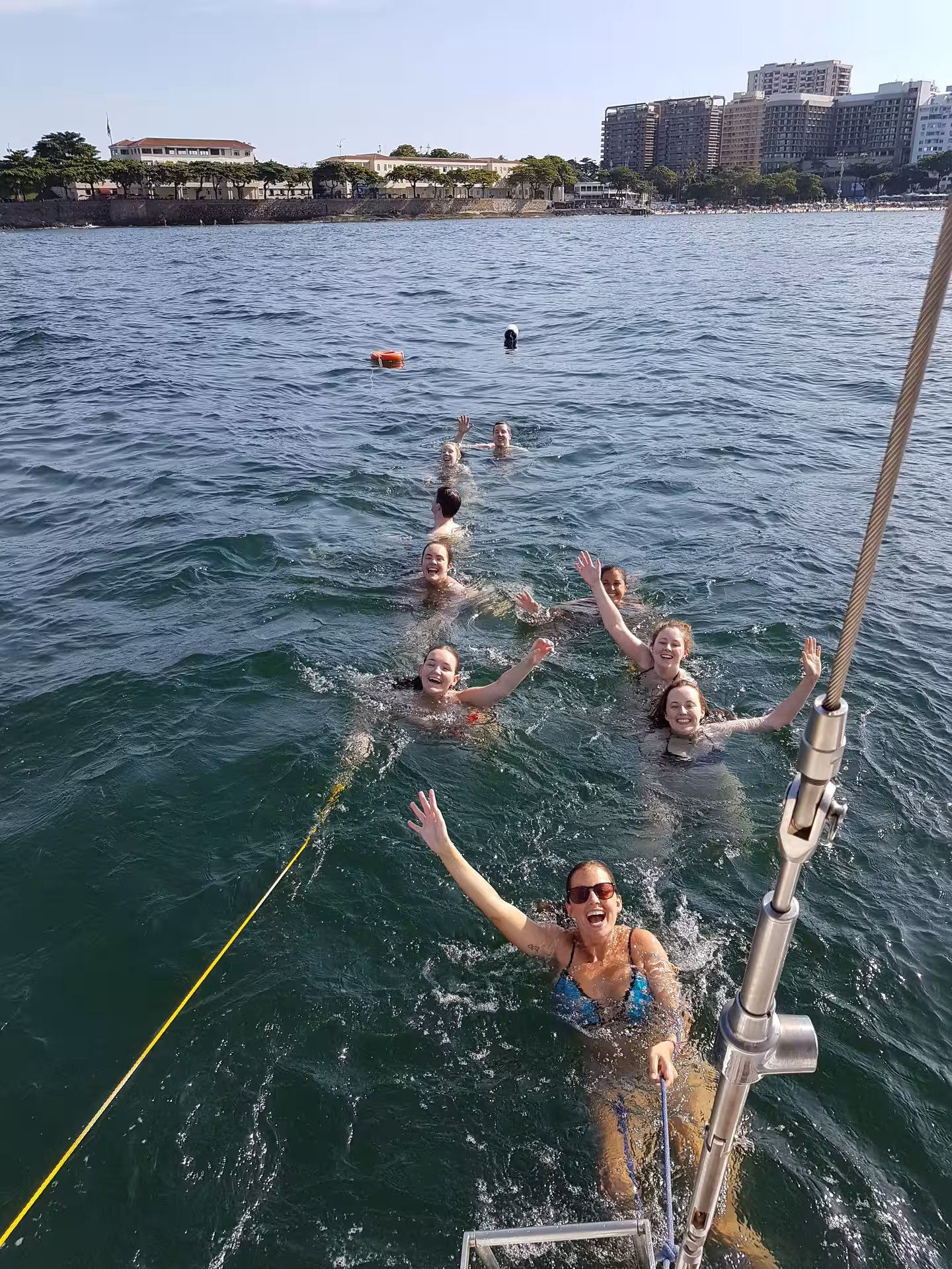 Group of friends joyfully swimming near a sailboat on a private Makani Maikai sailing tour in clear blue waters.