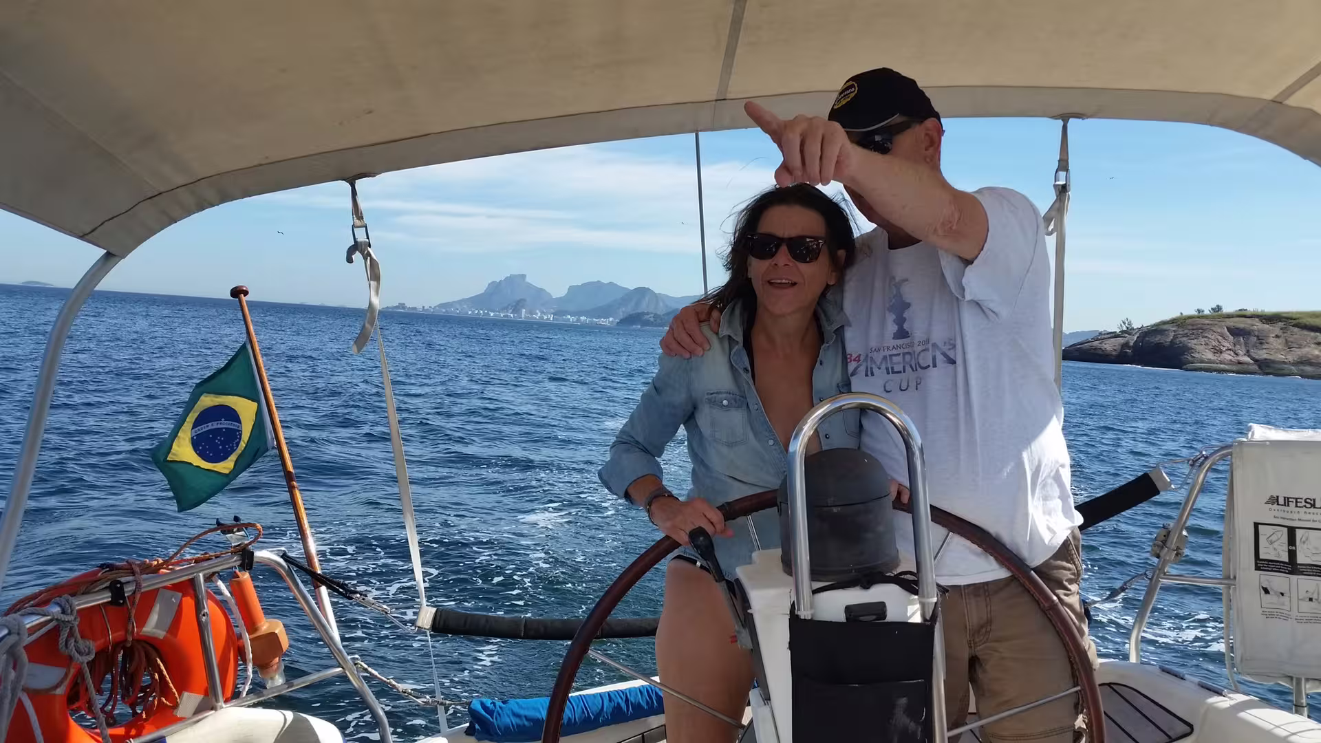 Couple steering the MAKANI MAIKAI sailboat on a serene ocean tour with scenic Brazilian coastline views.