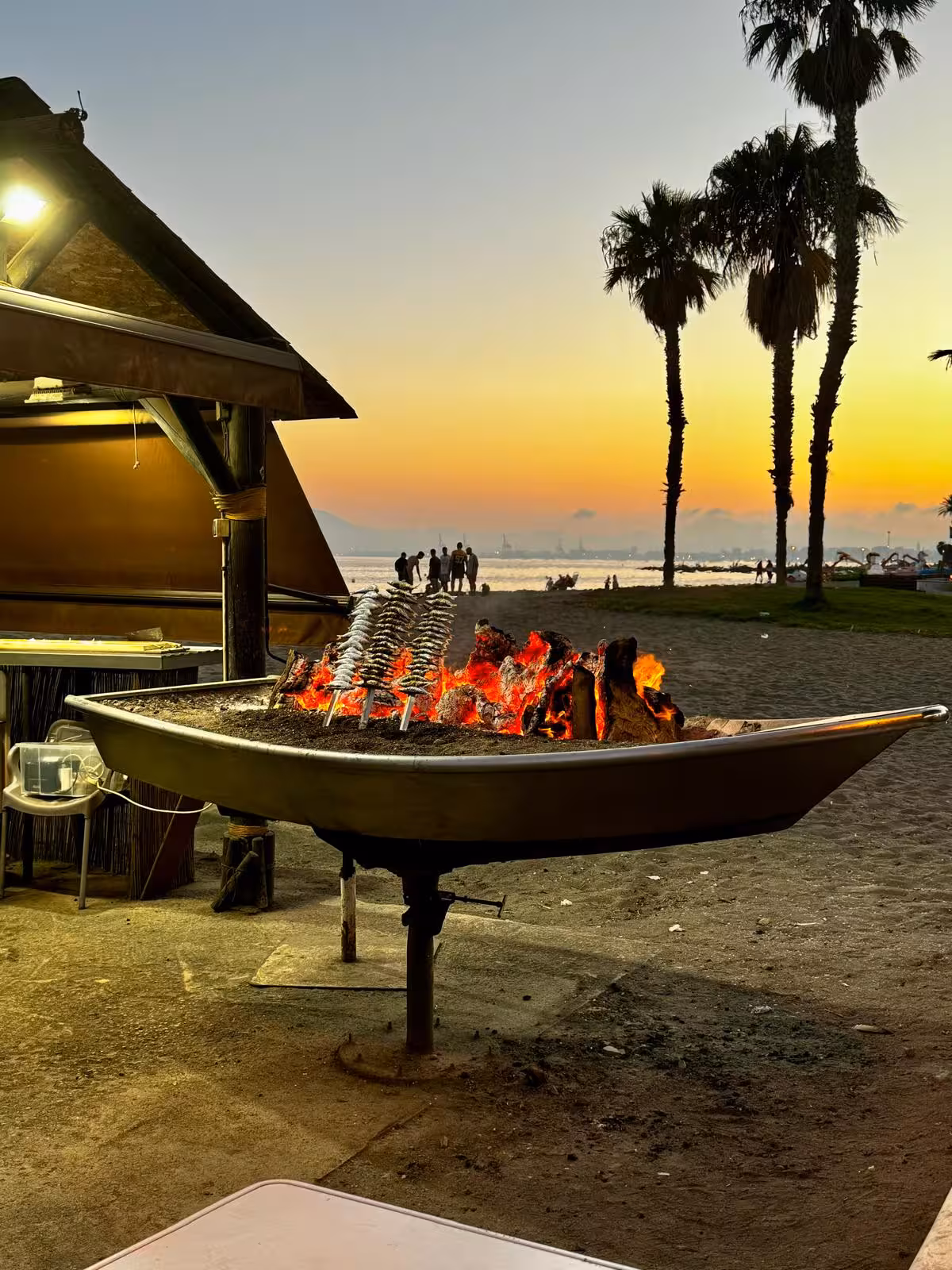 Outdoor barbecue with espetos, glowing coals on the El Palo beach during a Málaga sunset.