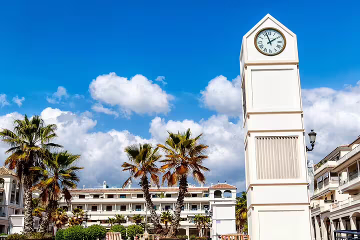 White clock tower with Roman numerals surrounded by palm trees in a sunny Mediterranean plaza