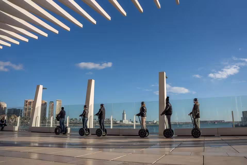 Segway riders explore Malaga's waterfront promenade under clear blue skies, with city views in the background.