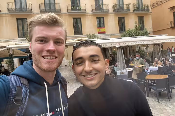 Two smiling men taking a selfie in Malaga's historic city center plaza with Spanish flag and taberna restaurant behind them
