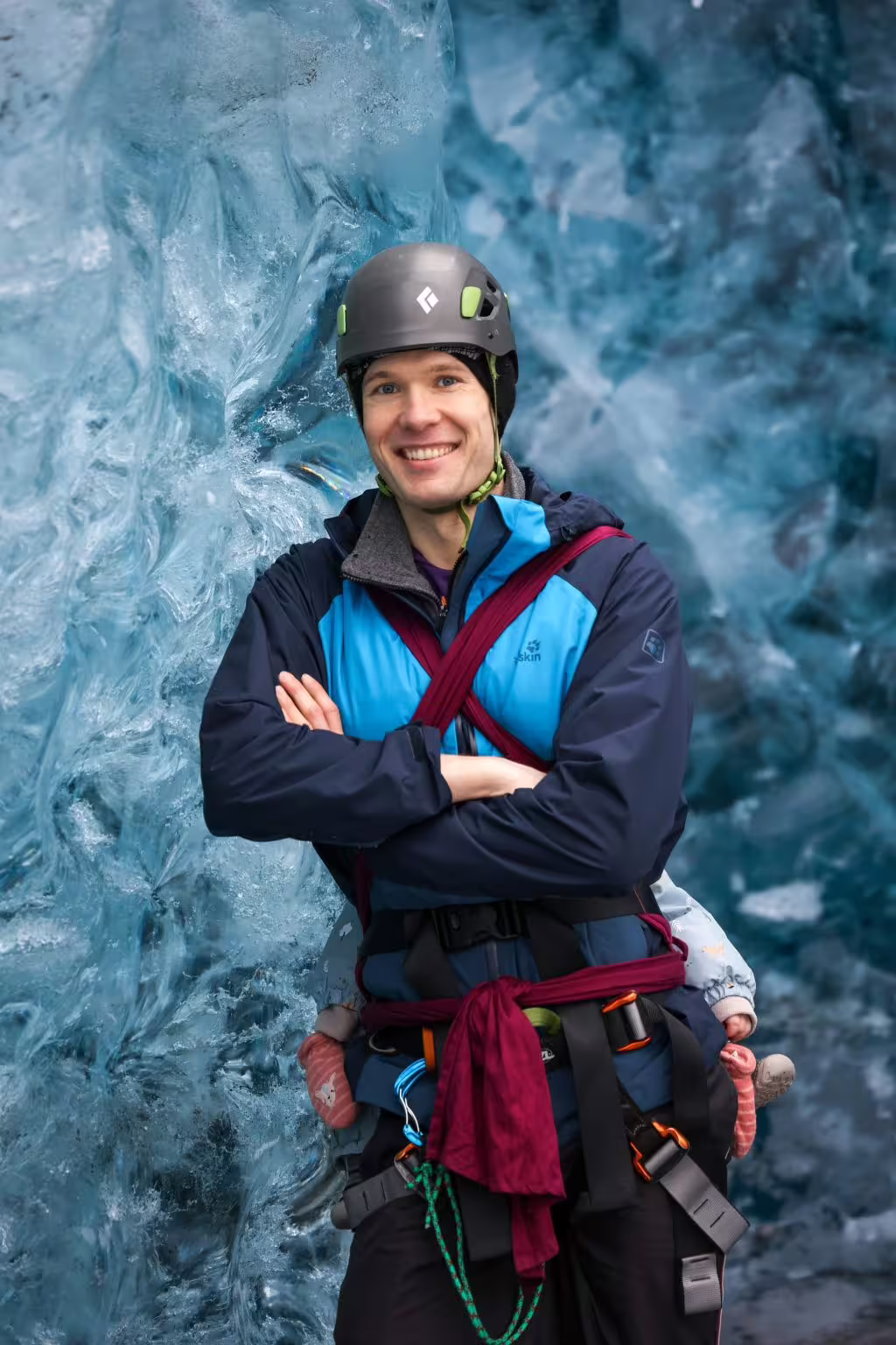 Smiling man in climbing helmet and harness posing confidently against a vivid blue glacial ice cave wall.