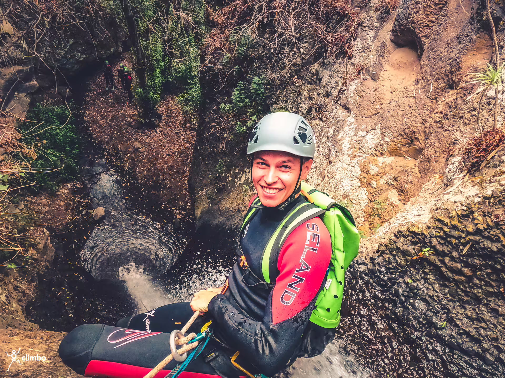 Smiling man rappels down a rocky waterfall in the rainforest, equipped with helmet and safety gear for a thrilling canyoning experience.