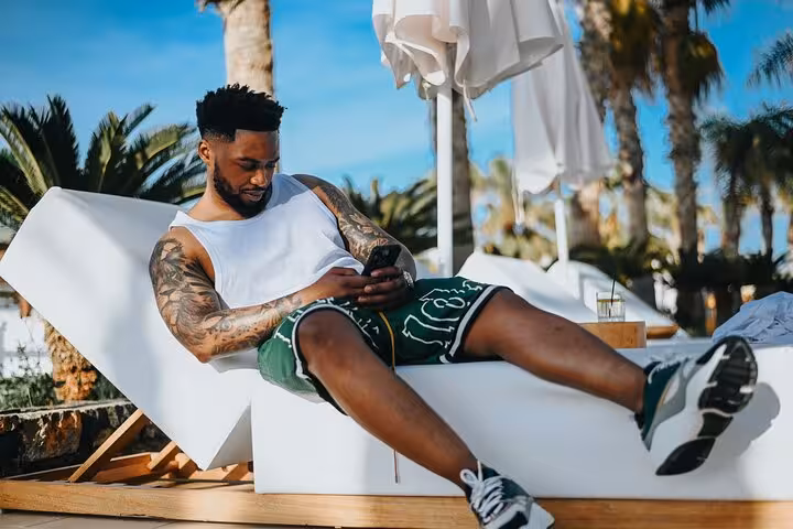 Man relaxing on a sun lounger, checking phone by the poolside under palm trees, Anisaras private photoshoot setting.