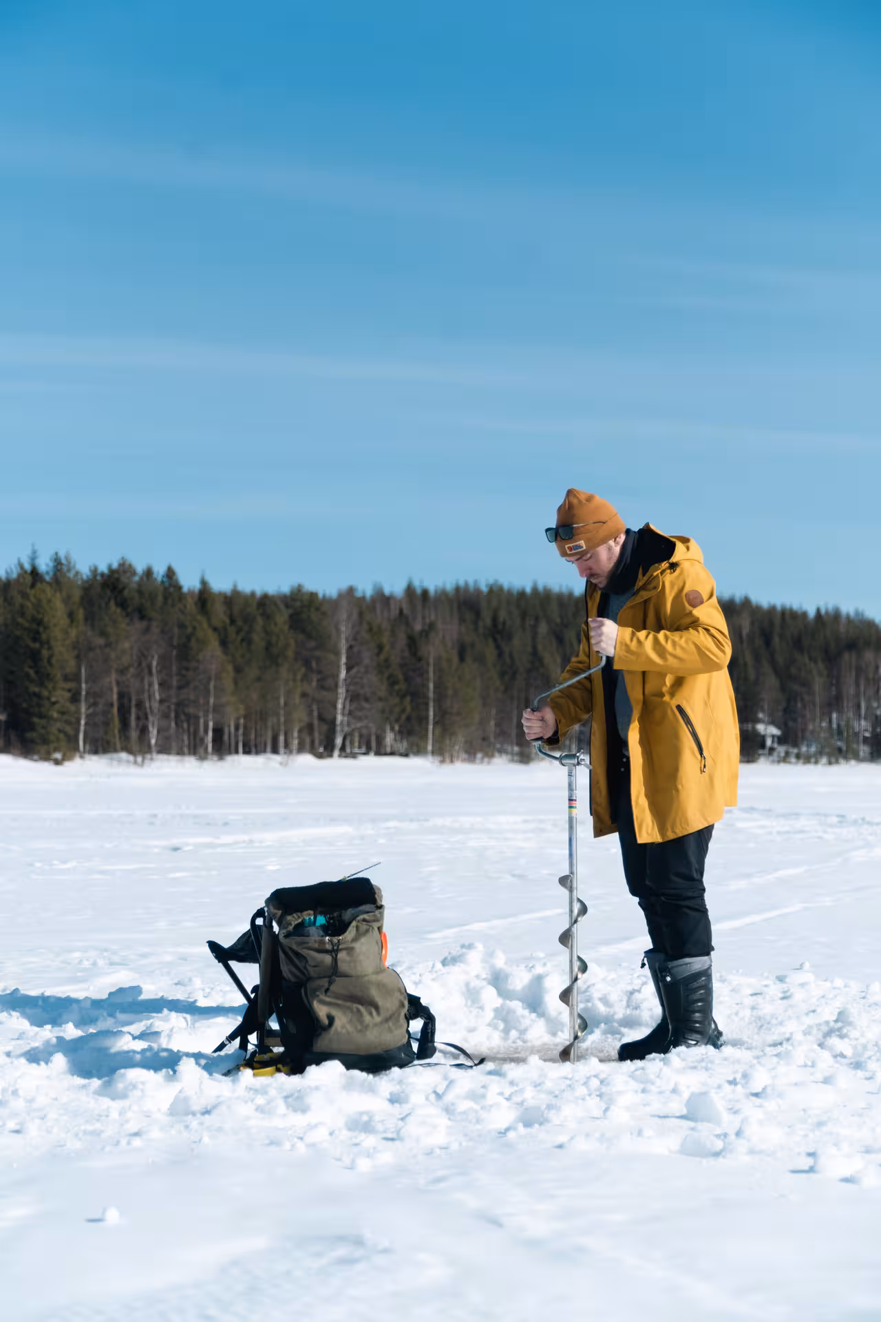 Man in yellow jacket drilling ice hole, preparing for local ice fishing adventure on a sunny winter lake.