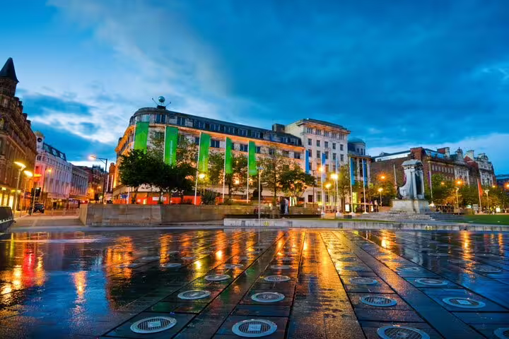 Wet Manchester city square at night with illuminated buildings and reflective paving stones