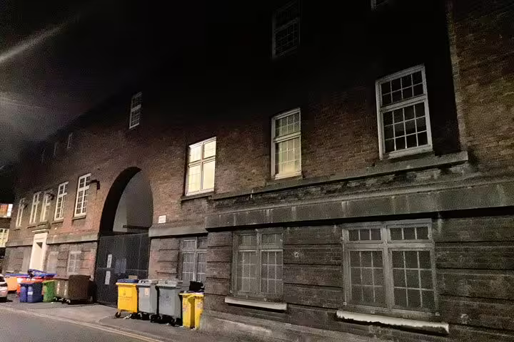 Dark brick building at night with arched gateway and yellow wheelie bins on a Manchester street