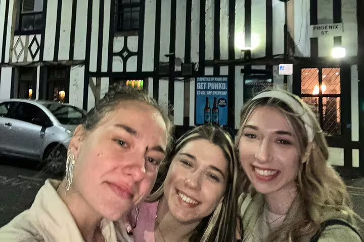 Three smiling women taking a selfie at night on a historic Manchester street with Tudor-style building behind them