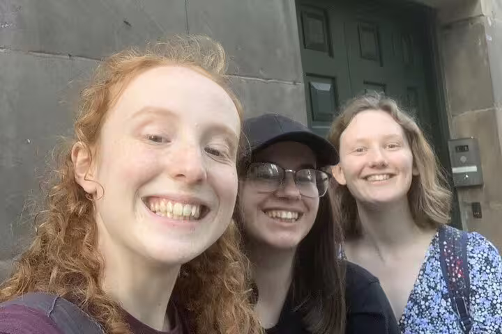 Three smiling young women taking a selfie outside a historic stone building in Manchester