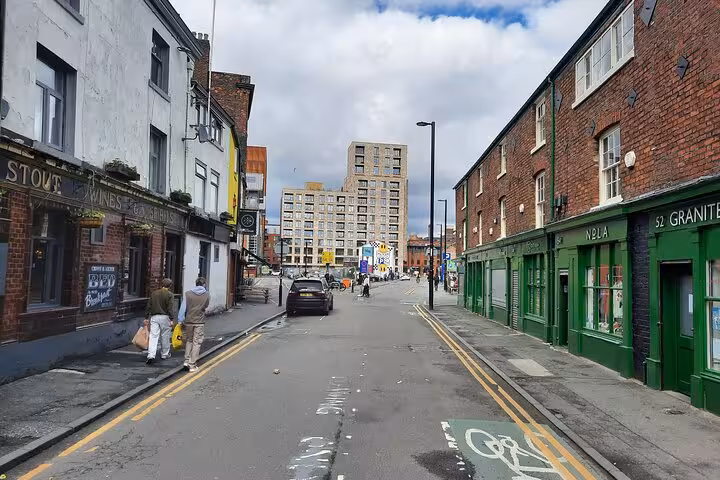 Manchester street with green-fronted shops, brick buildings, and a car on a quiet urban road