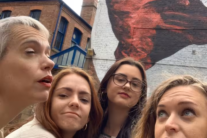 Four women posing for a selfie in front of a large red street art mural on a brick wall in Manchester