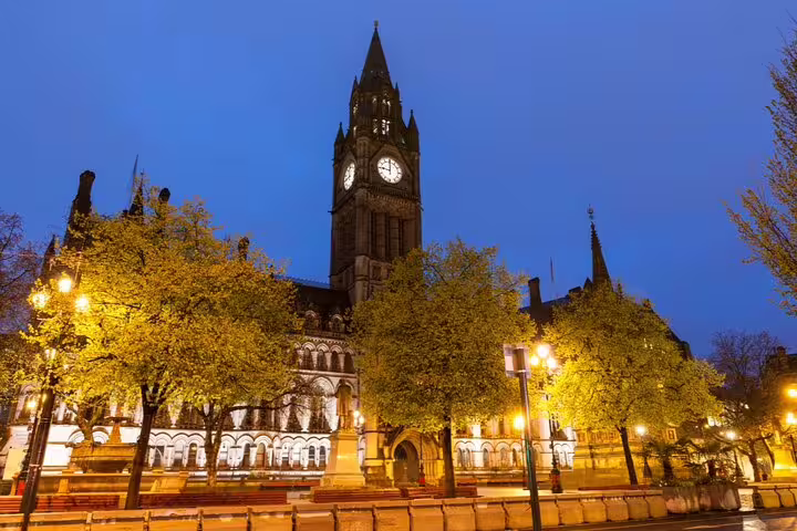 Manchester Town Hall clock tower illuminated at night with glowing street lamps and spring trees