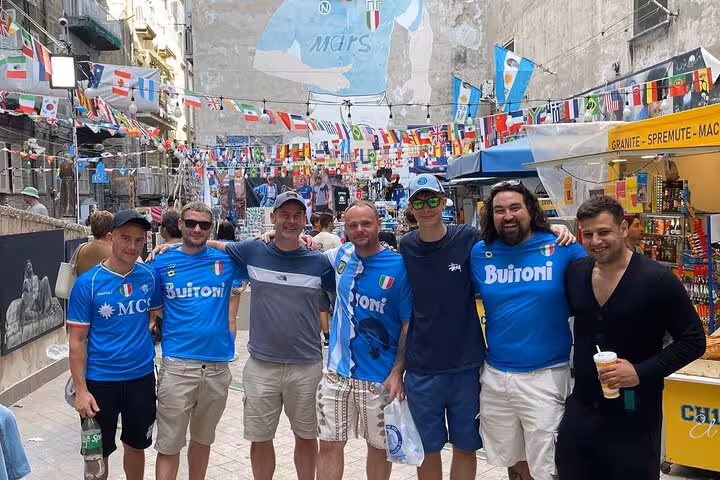 Fans in Napoli shirts on a lively Naples alley under flags, part of the Maradona legacy football and culture tour