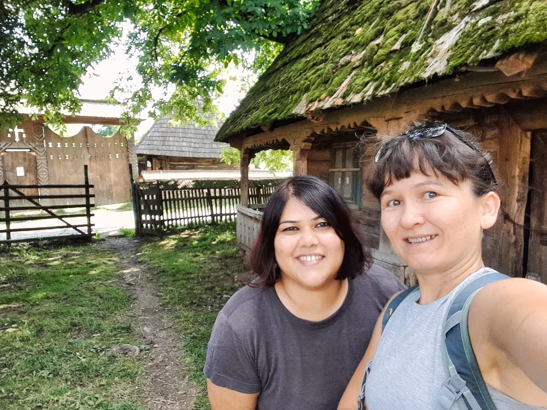 Two women smiling in selfie beside a moss-roofed traditional wooden house in Maramures, Romania