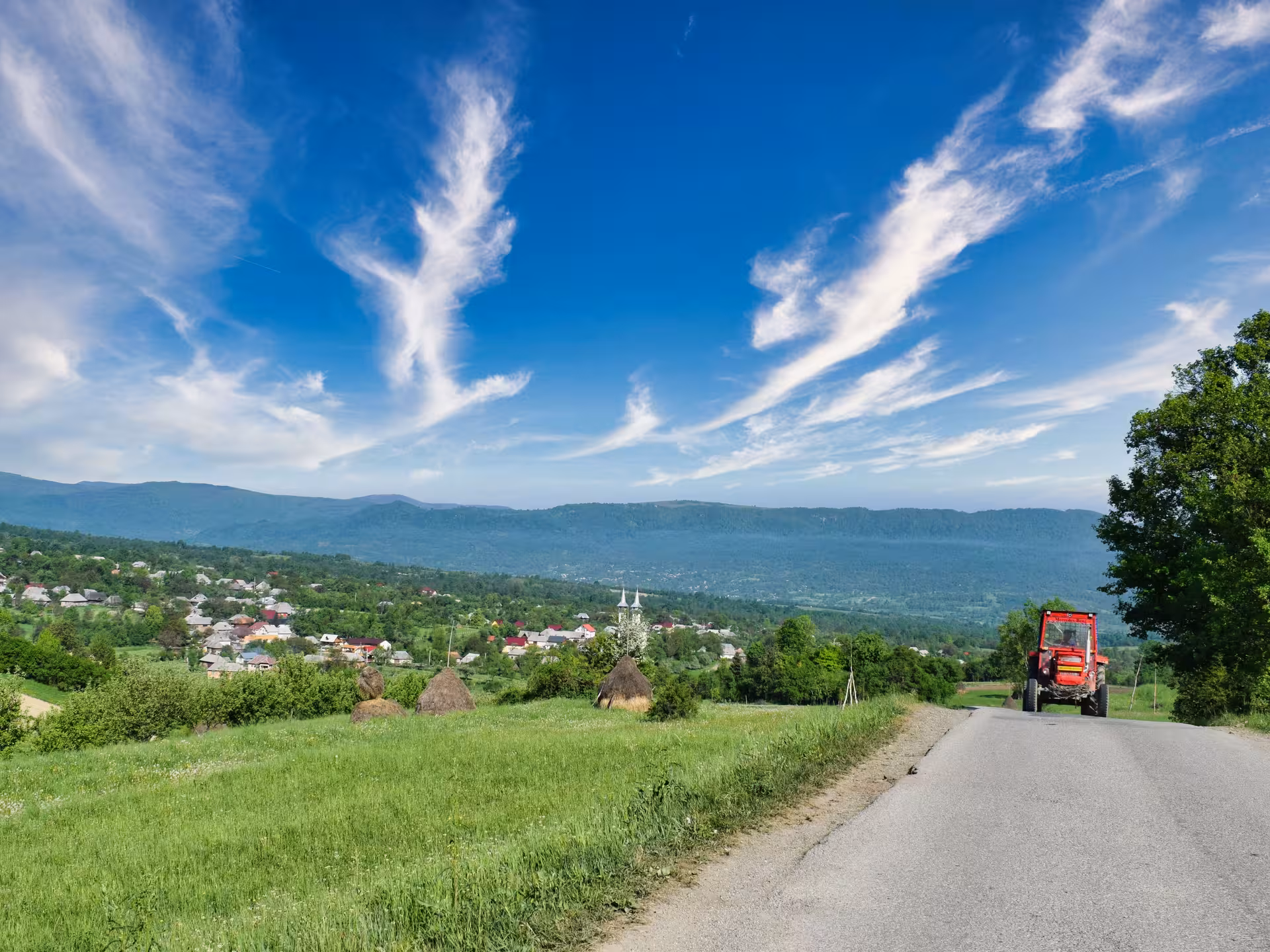 Red tractor on rural road with hay stacks, village, and mountain landscape in Maramures Romania
