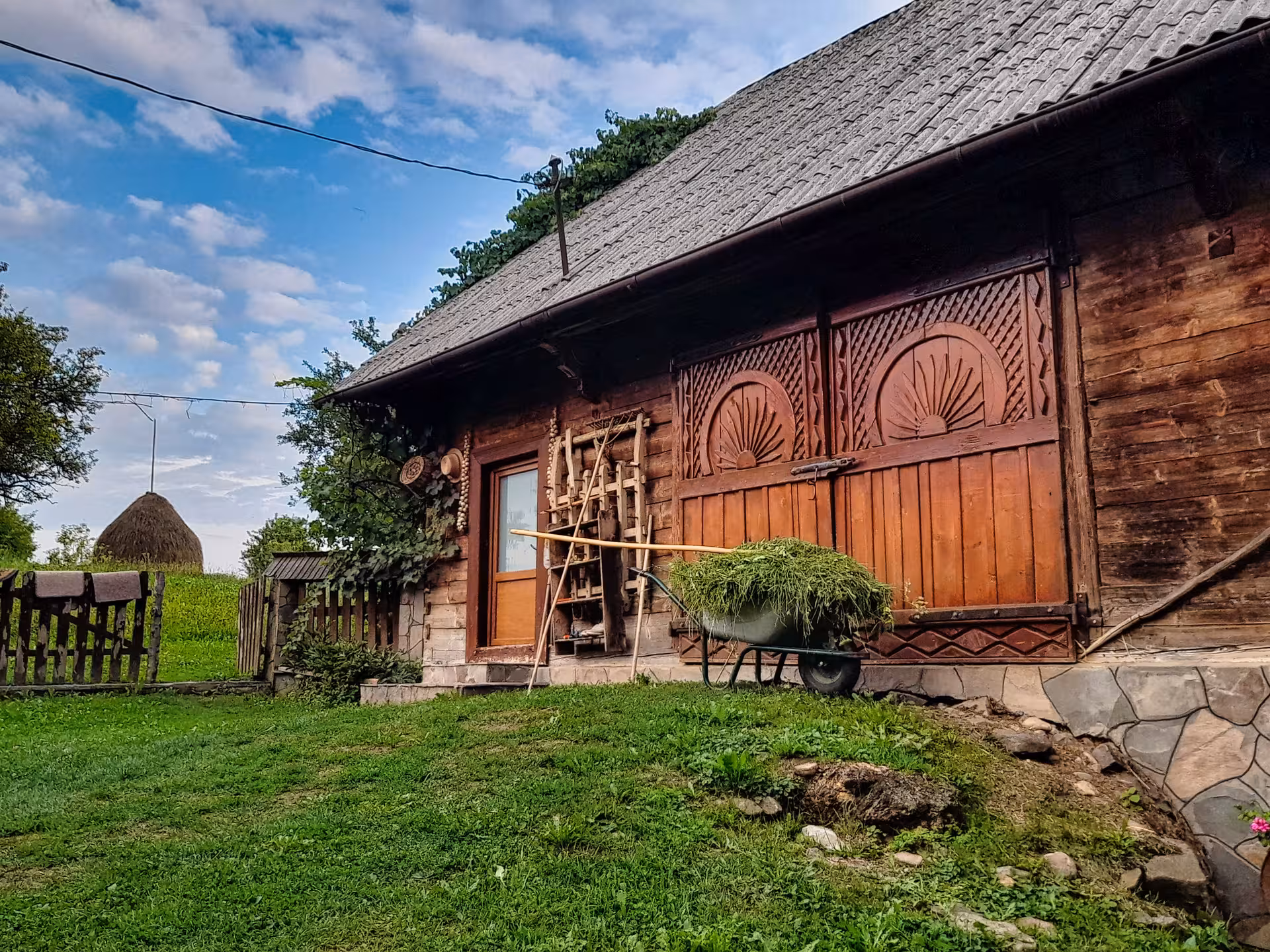 Traditional carved wooden farmhouse with wheelbarrow of hay and haystack in rural Maramures, Romania