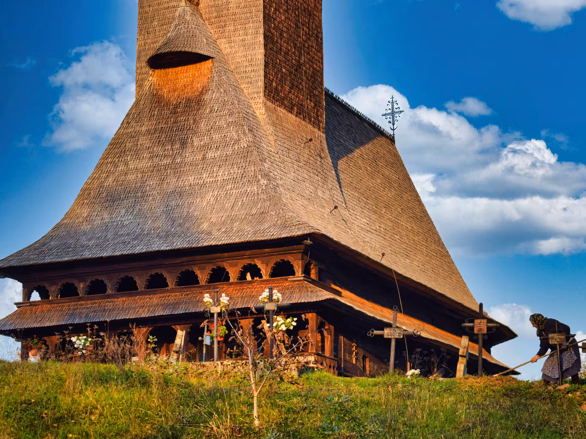 Tall wooden UNESCO church with shingled roof and ornate porch in Maramures, Romania, woman raking nearby