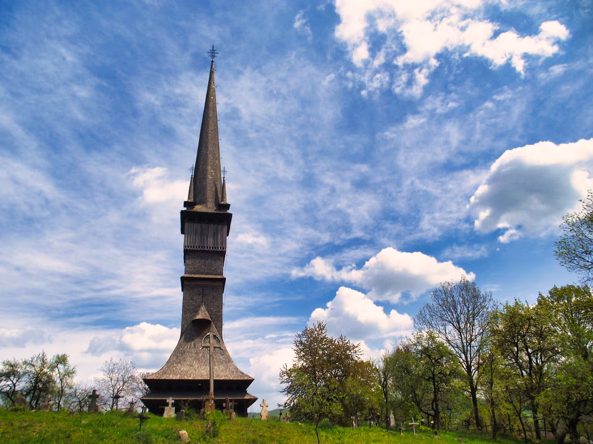 Tall wooden UNESCO church with soaring spire on green hill surrounded by trees and gravestones, Maramures Romania