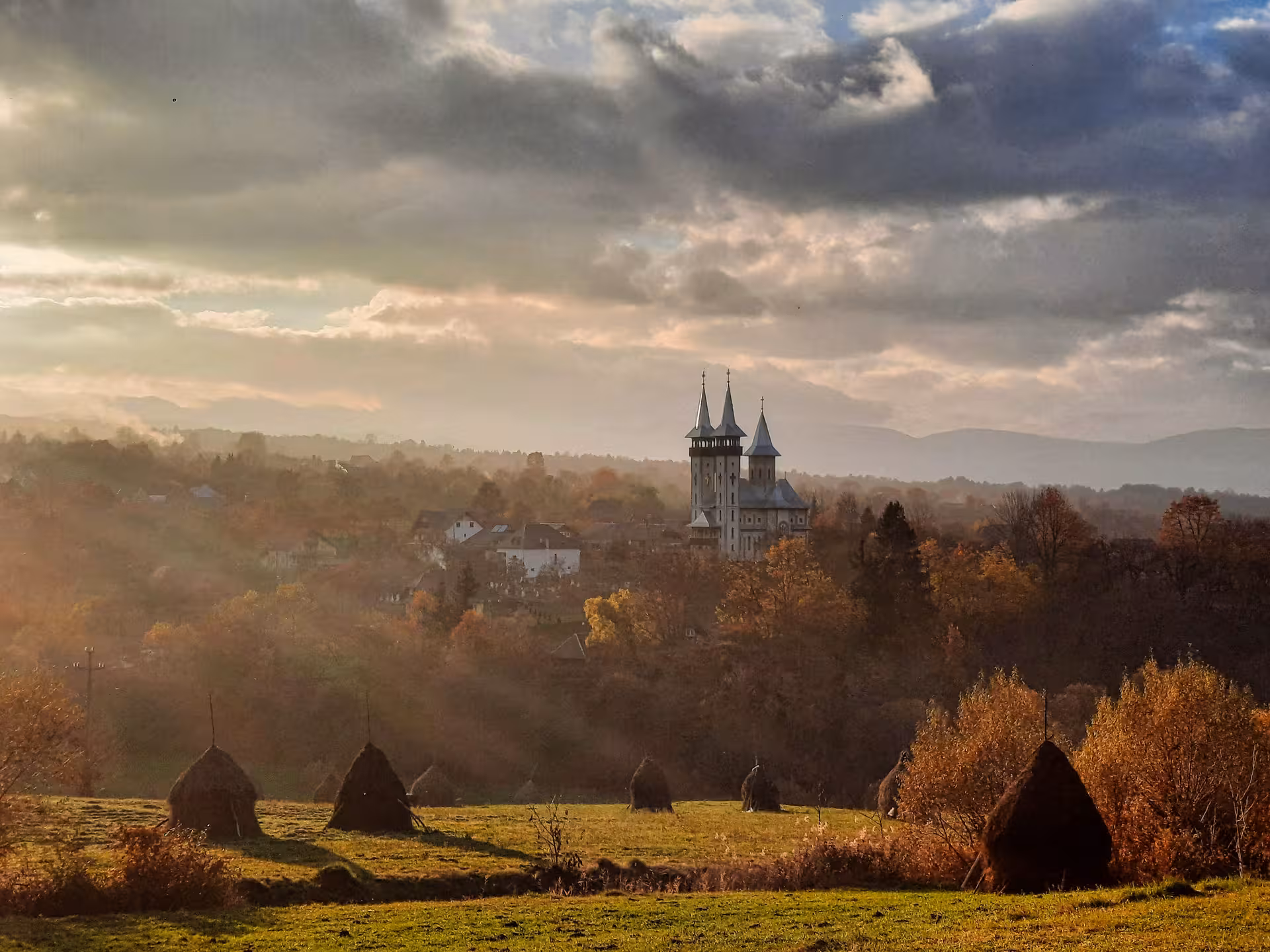 Scenic Maramures rural village with twin-tower church rising above autumn forest and traditional haystacks in golden light