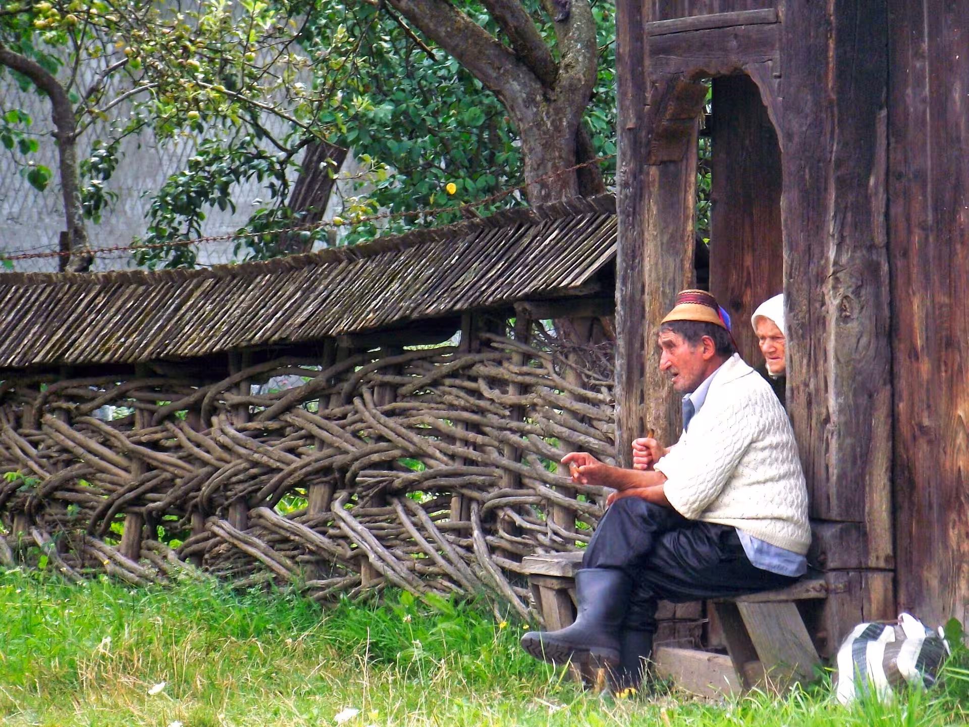 Elderly man in traditional cap sitting by carved wooden gate in Maramures village, Romania